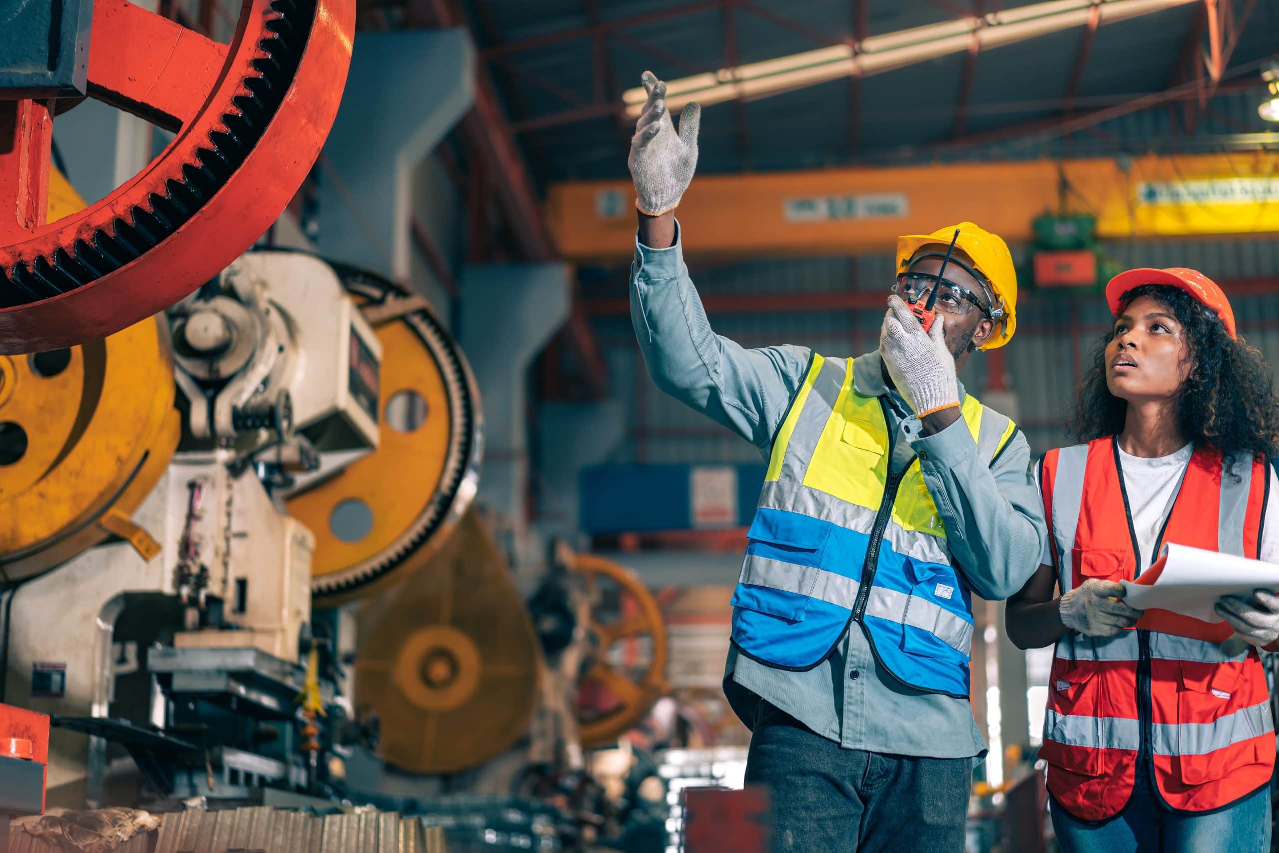 Two professional engineer/worker/technician use clipboard discuss work, walk in steel metal manufacture factory plant industry. Black African American man and woman wear hard hat check quality machine
